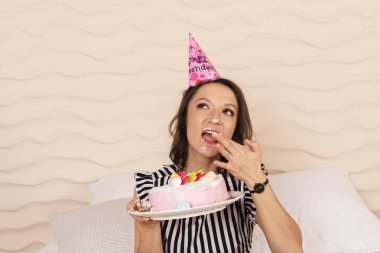 Güzel esmer kadın doğum gününü kutluyor, çizgili kısa bir elbise ve doğum günü şapkası giyiyor, yatağa oturuyor ve pembe pastadan bir parça yiyor. Yüzüne pasta bulaştırdı..