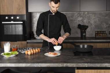 Mutfakta siyah üniformalı bir aşçı yemek pişirmek için malzemelerle hazırlandı. Şef kahvaltıda pastırmalı omlet yapıyor. Bir kaseye yumurta kırıyor..