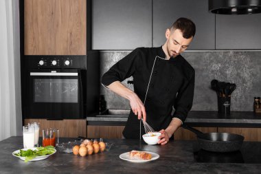 Mutfakta siyah üniformalı bir aşçı yemek pişirmek için malzemelerle hazırlandı. Şef kahvaltıda pastırmalı omlet yapıyor, kasede yumurta çırpıyor..