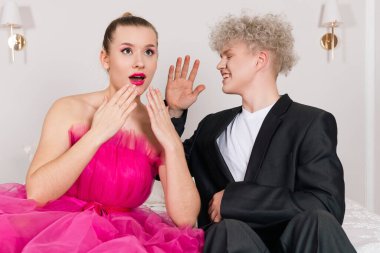 Bir Cazibeli Kız ve bebek gibi görünen bir adam, yatakta oturuyor. Uzun pembe elbiseli ve siyah takım elbiseli oyuncak bebek çifti. Dedikoducu ve bebek gibi hareketlerle kıkırdıyorlar..