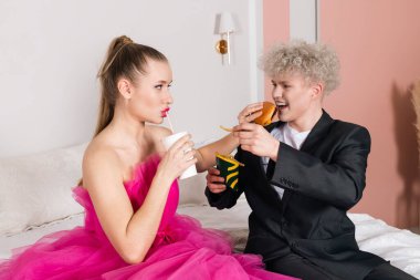 Çekici bir adam ve bebek gibi görünen bir kız, yatakta oturuyorlar. Pembe elbiseli ve siyah takım elbiseli bir bebek gibi davranıyor. Fast food, hamburger, patates kızartması ve soda. Sağlıksız diyet. oyuncak bebek gibi hareketler.