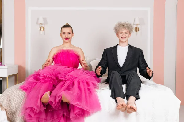 A Glamour Girl and a man looking like a doll, sitting on a bed. Doll couple dressed in long pink dress and black suit.