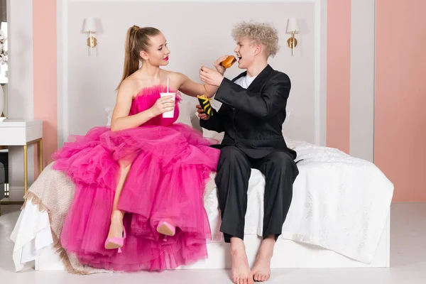 グラマーの男と女の子は人形のように見え ベッドの上に座っています ピンクのドレスと黒のスーツの人形になるふりをする ファーストフード ハンバーガー フライドポテト ソーダ 不健康な食事 人形のような動き — ストック写真