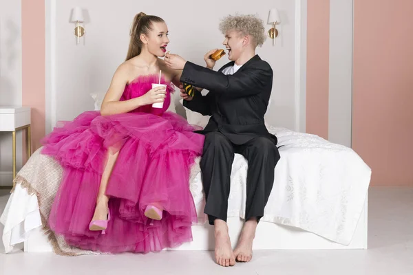 グラマーの男と女の子は人形のように見え ベッドの上に座っています ピンクのドレスと黒のスーツの人形になるふりをする ファーストフード ハンバーガー フライドポテト ソーダ 不健康な食事 人形のような動き — ストック写真