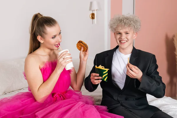 A Glamour man and a Girl looking like a doll, sitting on a bed. Pretending to be a doll in pink dress and black suit. fast food eating, burger, fries and soda. unhealthy diet. doll-like movements.