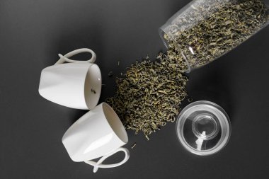 Beyaz bir fincan ya da bardak ve cam bir kavanoz dökülmüş yaprak çayıyla yan yana yatıyorlar. Düz görünüm, kopyalama alanı. koyu gri veya siyah arkaplanda izole edilmiş.