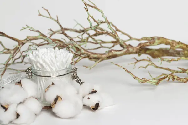 stock image glass jar with cotton ear buds decorated with a dried plant branches and cotton flowers lying on a white background.