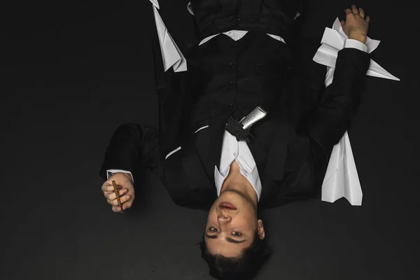 stock image Lying on a floor Young mafia member with a slicked back hair, dressed in black suit smoking a cigar, with a gun in his hand. defeated, dying shot Italian mafia gangster. isolated on black background.