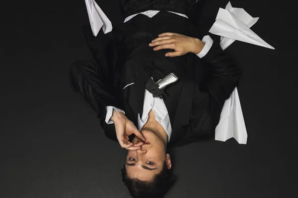 stock image Lying on a floor Young mafia member with a slicked back hair, dressed in black suit smoking a cigar, with a gun in his hand. defeated, dying shot Italian mafia gangster. isolated on black background.