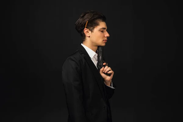 Portrait in profile. Hopeless and upset Young mafia member with a slicked back hair, in black suit, cigar behind the ear, pointing a gun to his chin. Italian mafia gangster. isolated on black.