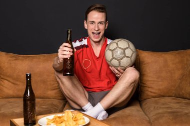 Kırmızı formalı bir futbol fanatiği TV 'de maç izliyor, kanepede oturuyor, bira içiyor ve cips yiyor..