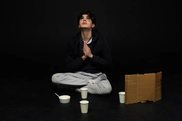 stock image A homeless young person with bruises under their eyes in dirty, tattered clothing, looks up and prays for a better life. Begging with an empty blank cardboard sign. Isolated on black background.