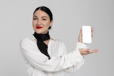 Çekici genç bir hostesin portresi. Çekici, güler yüzlü, şehvetli üniformalı hostes, boş beyaz şablon telefon görüntüsüyle gösterişli hareketler yapıyor. Beyaz arkaplanda izole.
