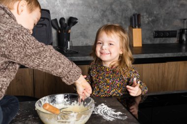 Küçük kardeşler modern bir mutfakta birlikte yemek pişiriyor, birbirlerine yardım ediyor ve eğleniyorlar, annem yokken çiğ hamur yiyorlar..