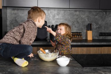 Küçük kardeşler modern bir mutfakta birlikte yemek pişiriyor, birbirlerine yardım ediyor ve eğleniyorlar, annem yokken çiğ hamur yiyorlar..