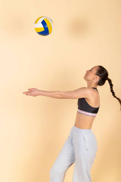 stock image Cute slim brunette girl in sportswear hits a volleyball. Young volleyball athlete spiking the ball. Isolated on pastel orange, peach background.
