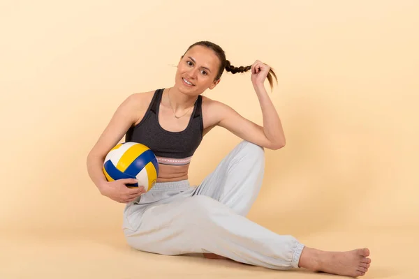 stock image Cute slim brunette girl in sportswear posing with a volleyball. Young athlete sits with a ball. Isolated on pastel orange, peach background.