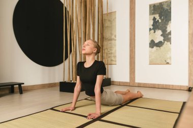 Sıska sarışın kız yoga yapıyor, egzersiz yapıyor, ısınıyor spor kıyafetleri giyiyor, pantolon ve bluz giyiyor, Japon tarzı bir stüdyoda..