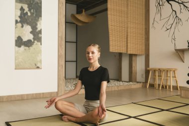 Sıska sarışın kız yoga yapıyor, egzersiz yapıyor, ısınıyor spor kıyafetleri giyiyor, pantolon ve bluz giyiyor, Japon tarzı bir stüdyoda..