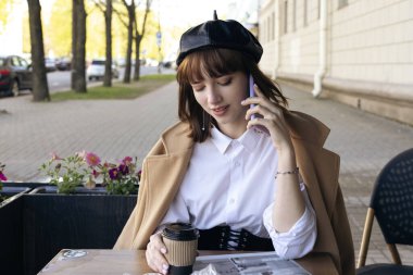 Saçları kısa saçlı, bereli, bej rengi klasik ceketli hoş bir esmer kahve, hamur işleri ve gazeteyle birlikte bir kafede oturuyor..