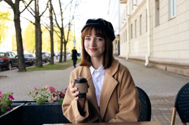 Kısa saçlı, bereli ve bej rengi klasik ceketli hoş bir esmer kahve, hamur işi ve gazeteyle birlikte bir kafe terasında oturuyor. Açık havada sabah, bahar.