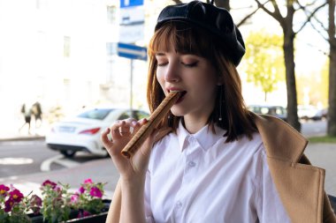 Kısa saçlı, bereli ve bej rengi klasik ceketli hoş bir esmer kahve, hamur işi ve gazeteyle birlikte bir kafe terasında oturuyor. Açık havada sabah, bahar.