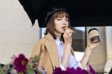 Kısa saçlı, bereli ve bej rengi klasik ceketli hoş bir esmer kahve, hamur işi ve gazeteyle birlikte bir kafe terasında oturuyor. Açık havada sabah, bahar.