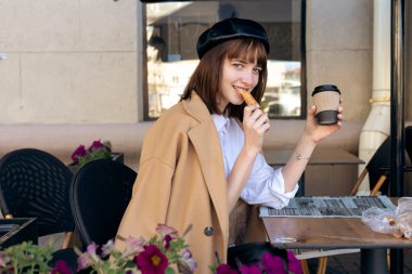 Kısa saçlı, bereli ve bej rengi klasik ceketli hoş bir esmer kahve, hamur işi ve gazeteyle birlikte bir kafe terasında oturuyor. Açık havada sabah, bahar.
