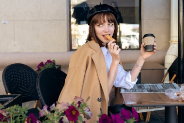 Kısa saçlı, bereli ve bej rengi klasik ceketli hoş bir esmer kahve, hamur işi ve gazeteyle birlikte bir kafe terasında oturuyor. Açık havada sabah, bahar.