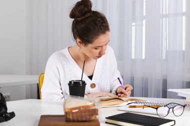 Genç kız öğrenci ödevini yapıyor, sınıfta çalışıyor. Masadaki dağınıklık, kitaplar, defterler, malzemeler. Şirin kız, saçları topuz gibi bağlanmış..