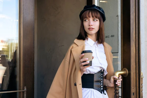 stock image A lovely young brunette with a bob haircut, wearing a beret and a beige classic coat, steps out of the coffee shop, opens the door of the building.