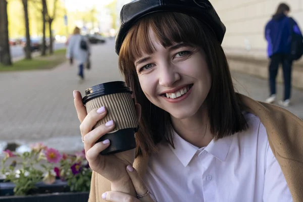 A lovely smiling young brunette with a bob haircut, wearing a beret and a beige classic coat, is sitting on a cafe terrace with a coffee, pastries and a newspaper. Outdoor morning, spring.