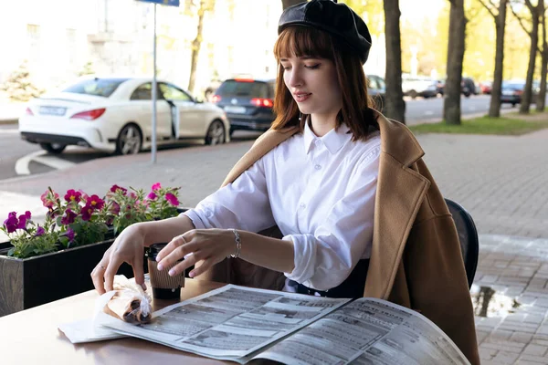 Kısa saçlı, bereli ve bej rengi klasik ceketli hoş bir esmer kahve, hamur işi ve gazete okuyan bir kafe terasında oturuyor. Açık havada sabah, bahar.