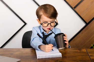 Yuvarlak gözlüklü, mavi gömlekli ve kravatlı küçük bir çocuk ofisteki masada oturmuş kağıt bardaktan içtiği not defterine not alıyordu. Zeki küçük çocuk, dahi çocuk..