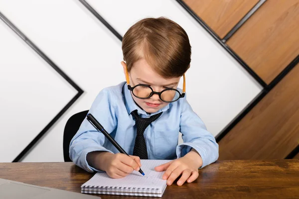 stock image Little boy in round eyeglasses, blue shirt and tie sitting at the desk in the office with a notepad. Smart little kid, Child prodigy.