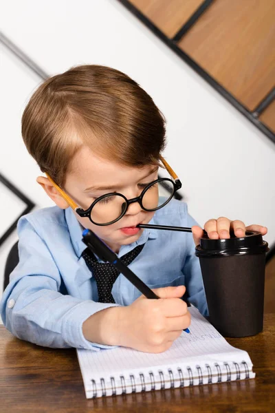 丸メガネの男の子 青いシャツとネクタイオフィスの机の上に座って 紙コップからメモ帳の飲み物でメモを取ります 賢い小さな子供 子供の神童 ロイヤリティフリーのストック写真