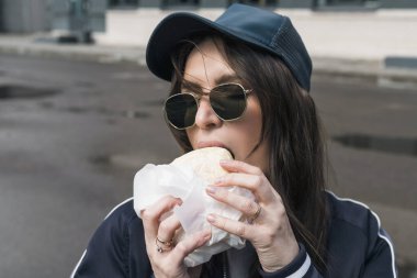 Spor takım elbiseli, güneş gözlüklü genç esmer kız sokakta fast food burgerini ısırıyor. Kafadan, yakın plan portre..