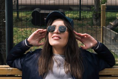 Mutlu gülümseyen esmer kız günlük spor kıyafetleri ve güneş gözlükleriyle sokakta, ahşap bir bankta oturuyor..