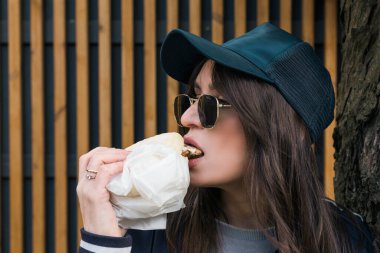 Spor takım elbiseli ve güneş gözlüklü esmer kız sokakta fast food burger yiyor..