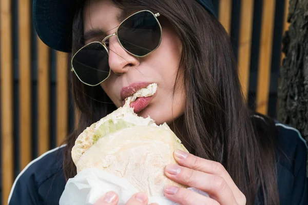 穿着休闲装和太阳镜的年轻黑发女孩在街上吃饭 在户外咬快餐汉堡 — 图库照片