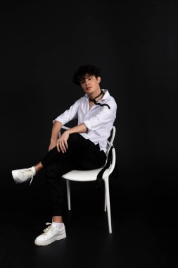 Boynunda siyah bir yılanla beyaz gömlekli genç bir adam sandalyede oturuyor. Siyah arkaplanda izole.