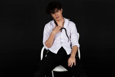 Boynunda siyah bir yılanla beyaz gömlekli genç bir adam sandalyede oturuyor. Siyah arkaplanda izole.