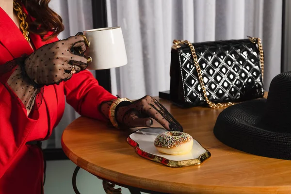 赤い服に巻き毛のエレガントなブルネットの女性 ポルカドットレース手袋は ドーナツとコーヒーを飲みながら 舗装カフェに座っています — ストック写真