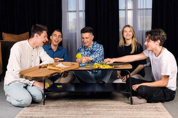 Five friends spending time together, having fun, playing table games and eating fast food and snacks. Small group of friend in a living room.