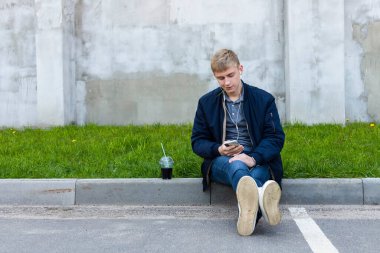 Sıcak bir bahar zamanı, gölgedeki kaldırımda oturan gündelik kıyafetli genç sarışın adam..