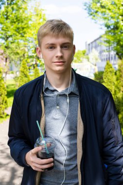 Sıradan kıyafetler ve kulaklıklar takan genç sarışın adam baharın ilk güneşli günlerinin tadını çıkarıyor. Plastik bardaktan kokteyl içiyor ve parkta yürüyor..