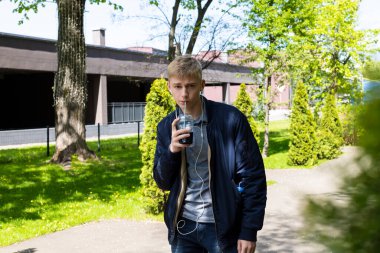 Sıradan kıyafetler ve kulaklıklar takan genç sarışın adam baharın ilk güneşli günlerinin tadını çıkarıyor. Plastik bardaktan kokteyl içiyor ve parkta yürüyor..