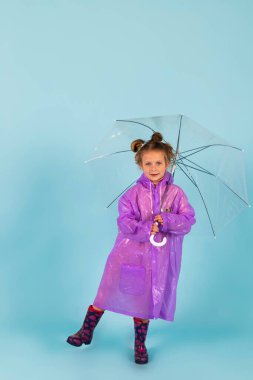 Mor yağmurluklu ve lastik çizmeli sevimli küçük bir kız şemsiye tutuyor. Yağmurlu bir hava için giyinmiş bir çocuk. Mavi arkaplanda izole.