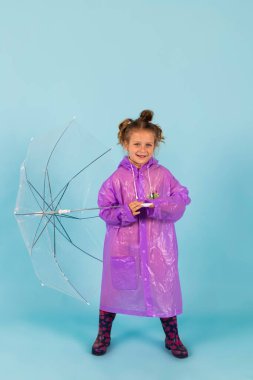 Mor yağmurluk ve lastik çizme giyen sevimli küçük kız temiz bir şemsiye tutuyor. Yağmurlu bir hava için giyinmiş bir çocuk. Mavi arkaplanda izole.