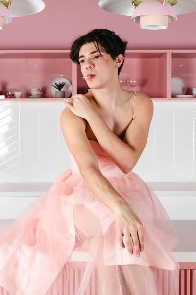 stock image Portrait of a tender gay in a pink dress with a lollipop. Glamour homosexual young guy with makeup posing in a girly outfit.